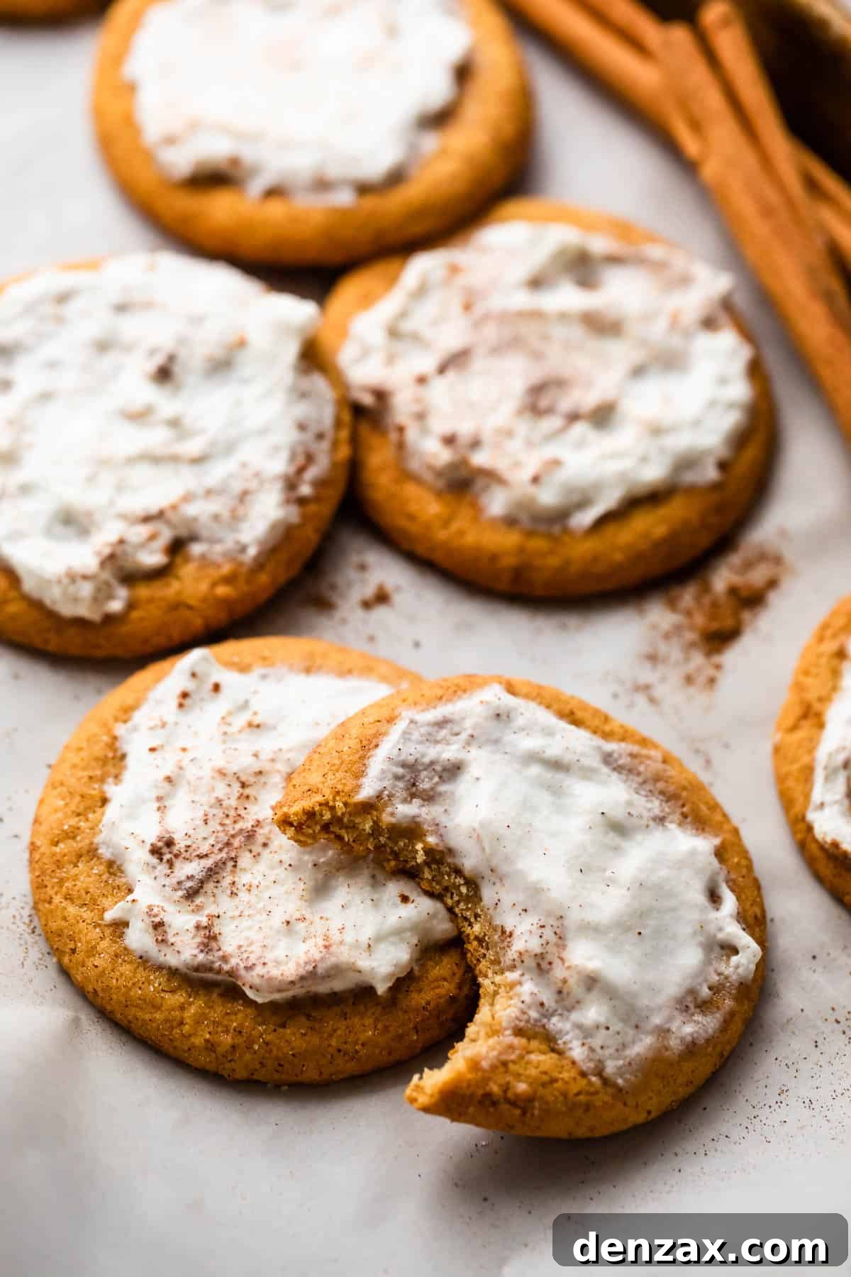 A charming scene of frosted pumpkin cookies, one with a bite taken out, surrounded by autumn cinnamon sticks and a rustic background, highlighting their cozy appeal and delicious texture.