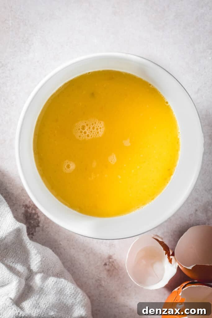 A white mixing bowl holds whisked eggs and chicken broth, forming the liquid component of the stuffing mixture, with discarded eggshells nearby on the kitchen counter.