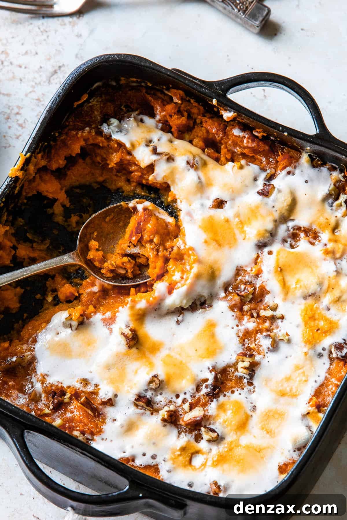 A vibrant sweet potato casserole with a spoon scooping out a generous portion, showcasing the golden-brown melted marshmallows and visible pecan pieces.