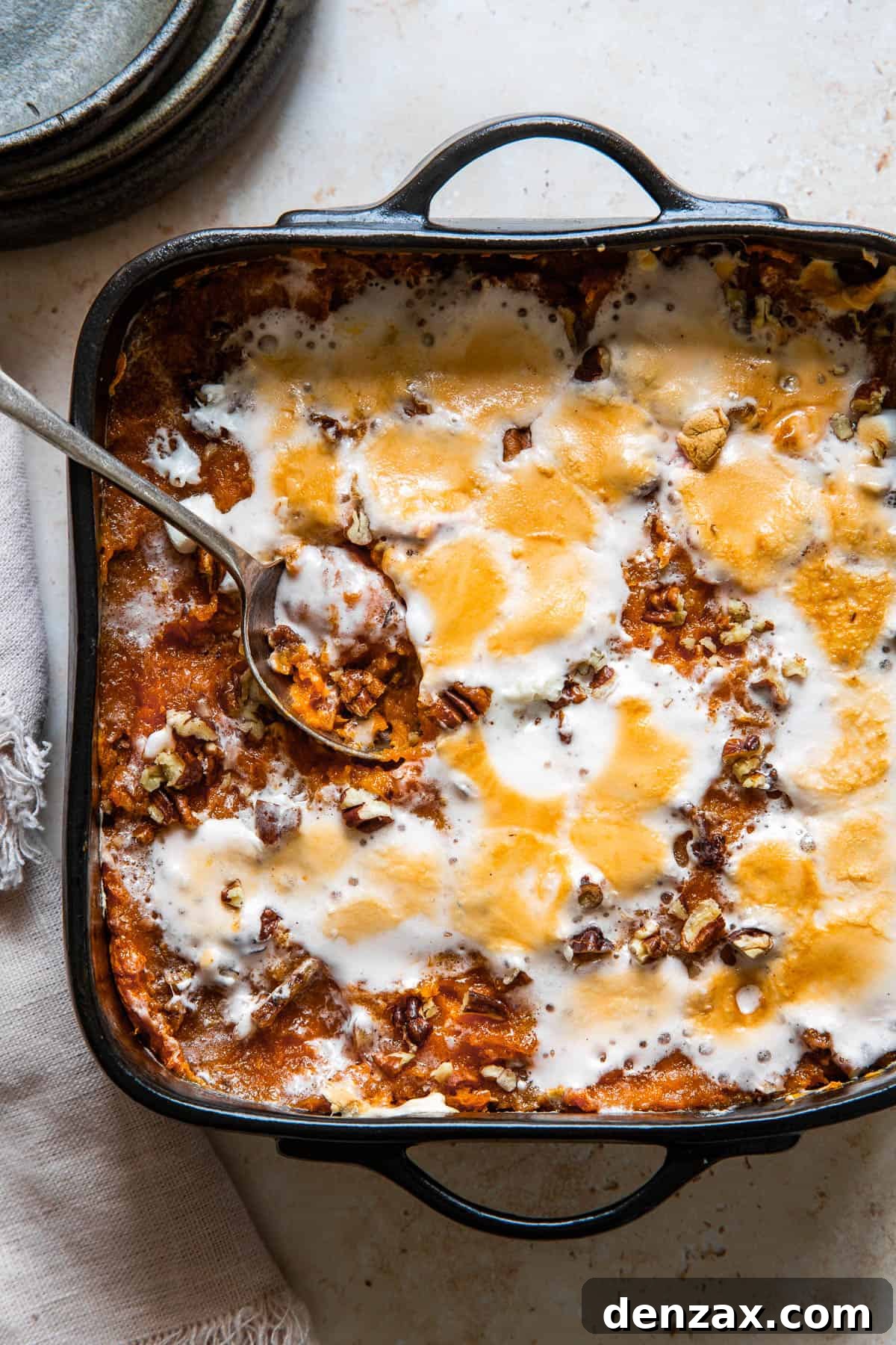 A serving spoon resting in a square black baking dish of golden-brown sweet potato casserole, topped with perfectly melted and toasted marshmallows and pecans.