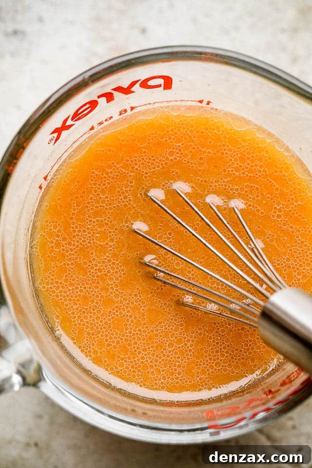 A close-up of a whisk stirring a homemade chicken stir fry sauce in a clear glass bowl, showcasing the rich, savory liquid ready to be added to the wok.