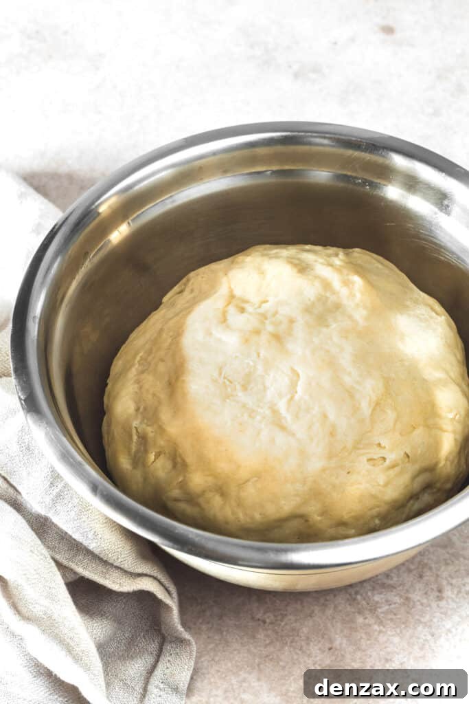 A stainless steel mixing bowl filled with a ball of soft, sticky dough, covered for rising.