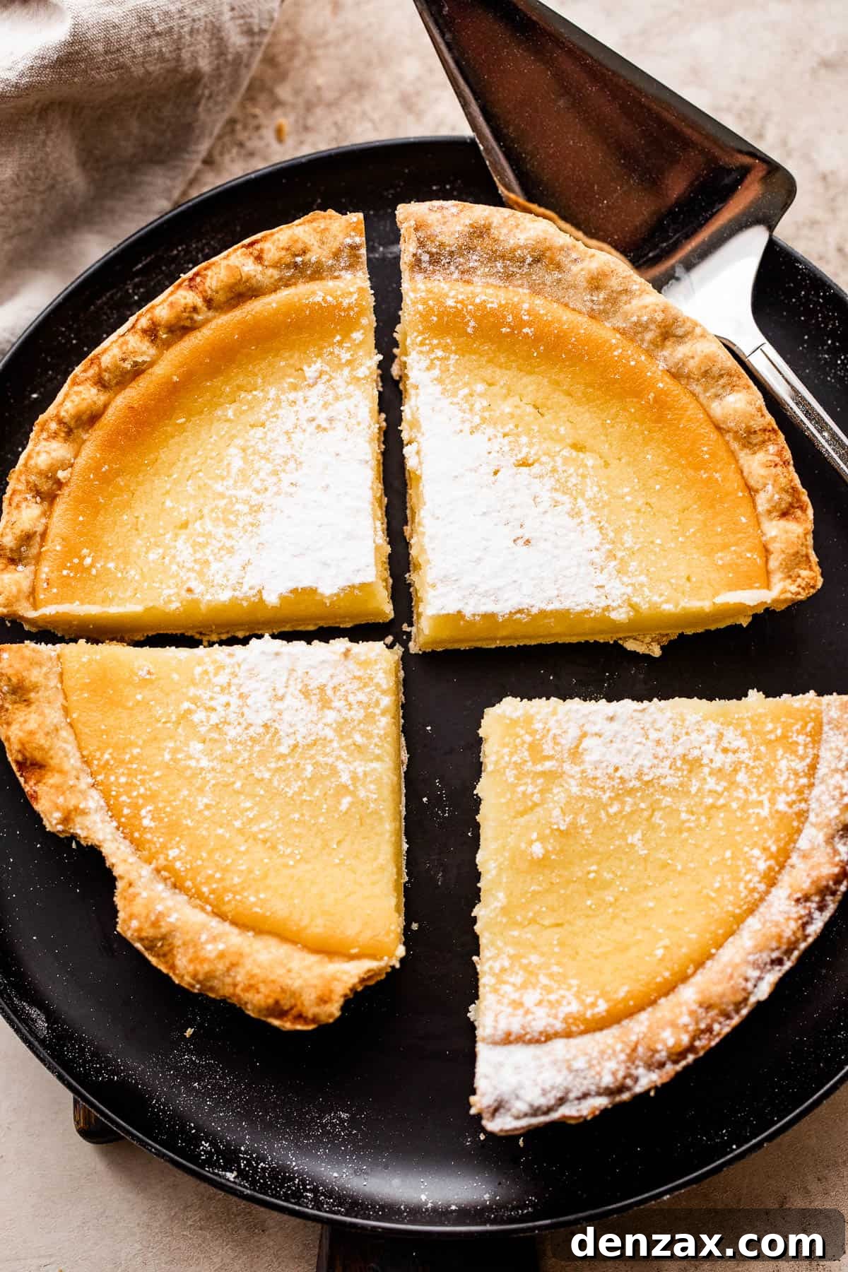 An overhead shot of a freshly baked Southern Buttermilk Pie, sliced into four generous portions and lightly dusted with powdered sugar, resting on an elegant black plate.