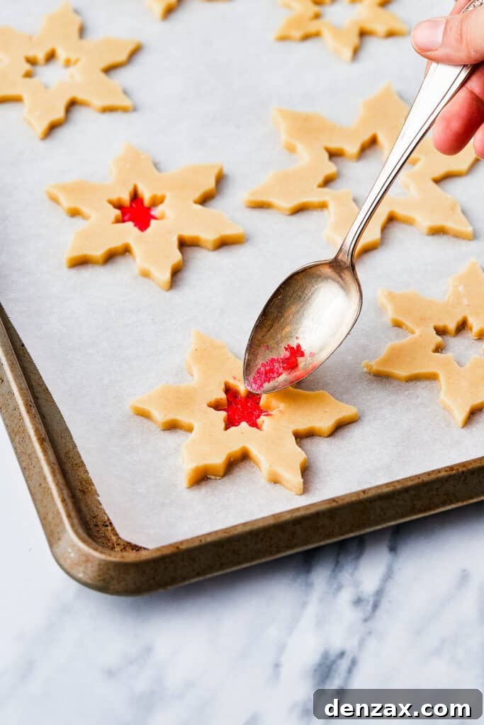 Cut out snowflake cookies with stars cut out of the center. A spoon is filling the centers with crushed red candy.