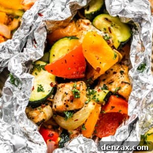 Overhead shot of an opened foil packet revealing perfectly cooked diced chicken, sliced zucchini, and sliced peppers, garnished with parsley and red pepper flakes.