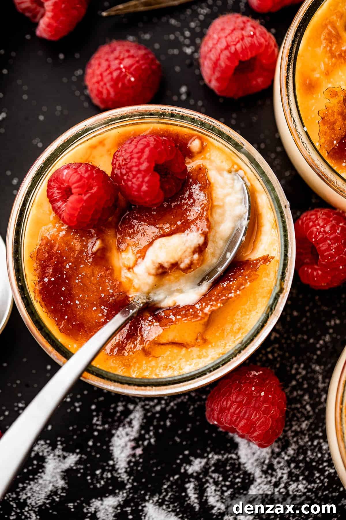 Overhead shot of a creamy Creme Brulee in a glass jar, adorned with two fresh raspberries, and a spoon gently dipped into the side, highlighting the golden caramelized sugar topping.