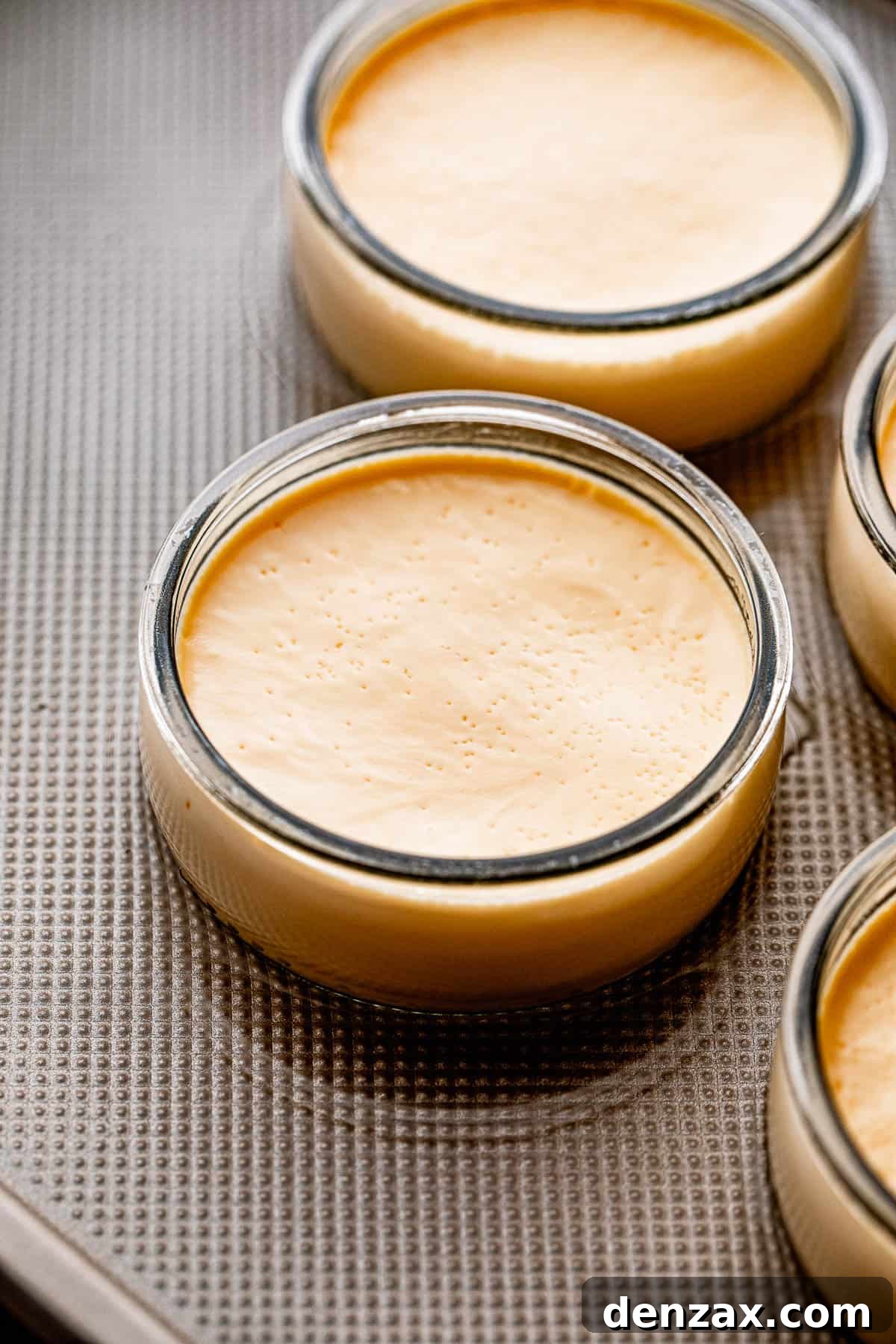 Side shot of two individual glass jars containing Creme Brulee, placed on a sheet pan filled with water, ready for baking in a bain-marie.