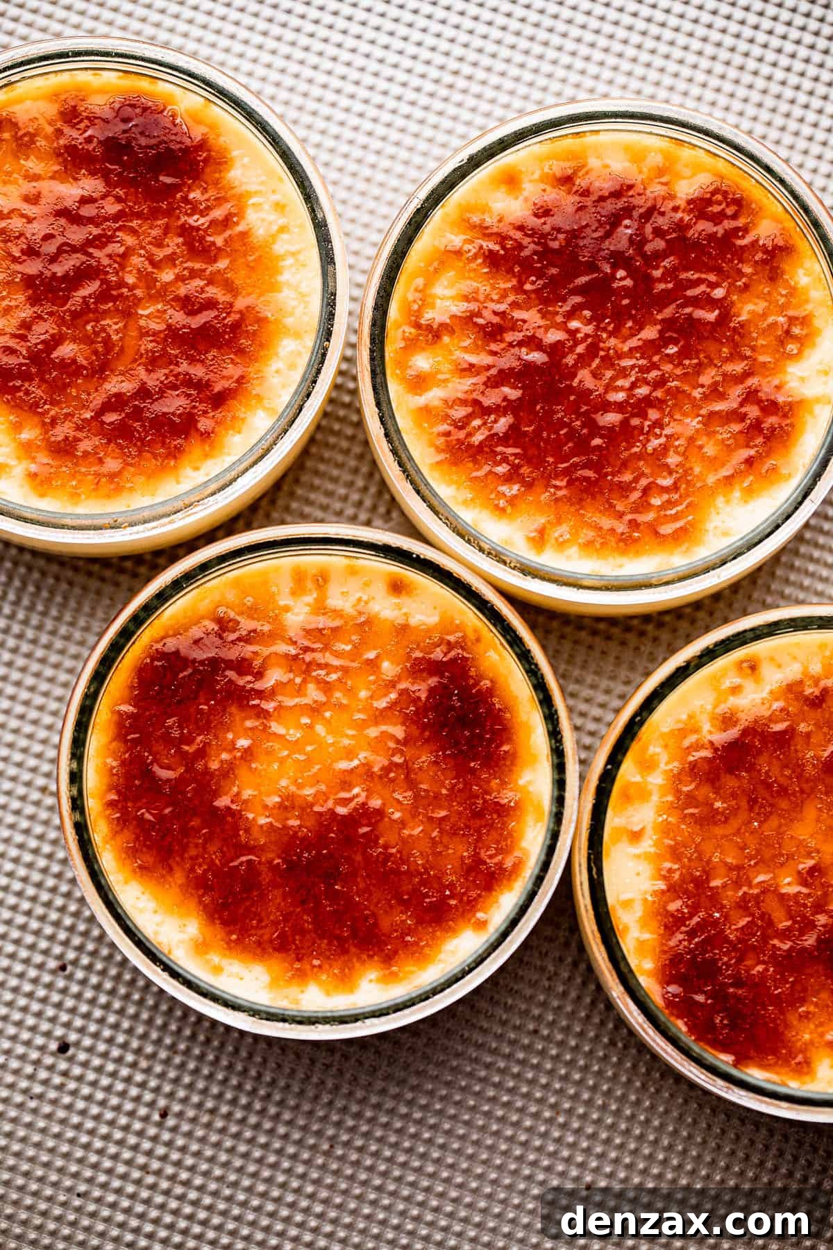 Overhead shot of four small jars, each containing smooth Creme Brulee custard, neatly arranged on a silver-colored background, showcasing the beautiful yellow hue of the dessert before caramelization.