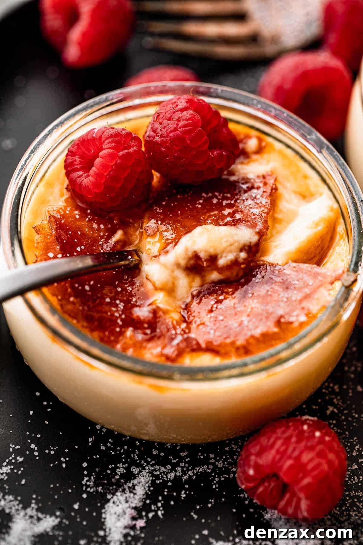 Side shot of a luxurious Creme Brulee in a glass jar, garnished with two fresh raspberries, and a spoon placed invitingly inside the jar.