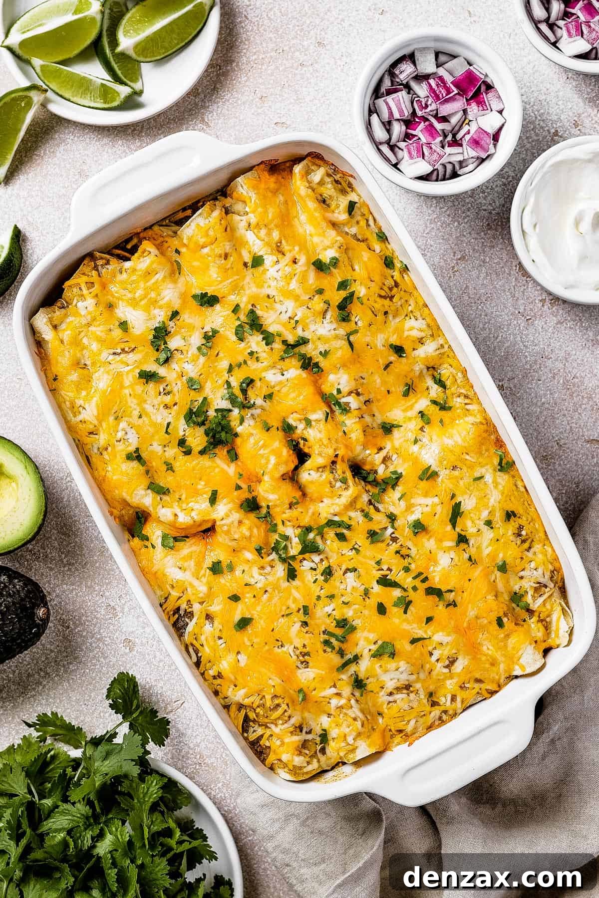 Overhead shot of freshly baked salsa verde chicken enchiladas in a white baking dish, garnished with fresh cilantro. Small bowls of diced red onion, sour cream, and lime wedges are set to the side.