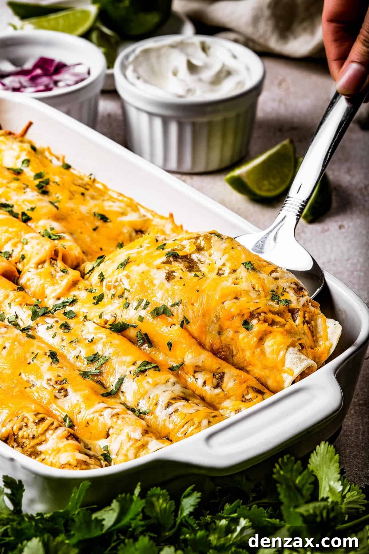 A silver serving spoon is lifting two golden-brown baked chicken enchiladas from a white ceramic baking dish, showing off the melted cheese and vibrant green sauce.