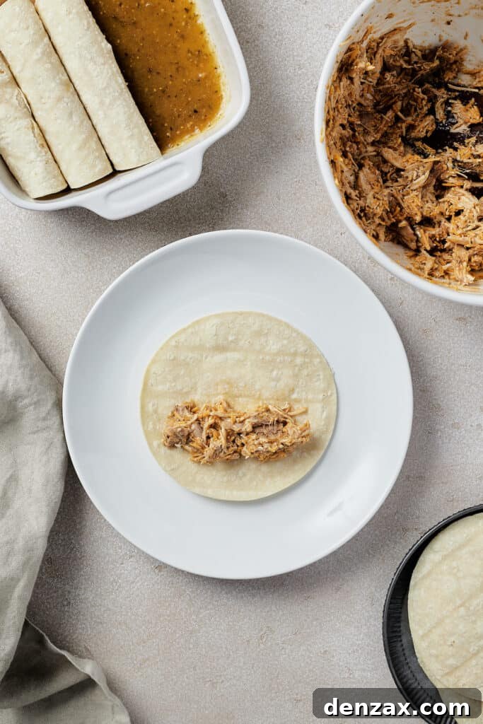 A small corn tortilla on a plate, with a spoonful of the chicken enchilada filling neatly arranged down its center, ready to be rolled. In the background, a baking dish of finished enchiladas, a bowl of more filling, and a stack of tortillas are visible.