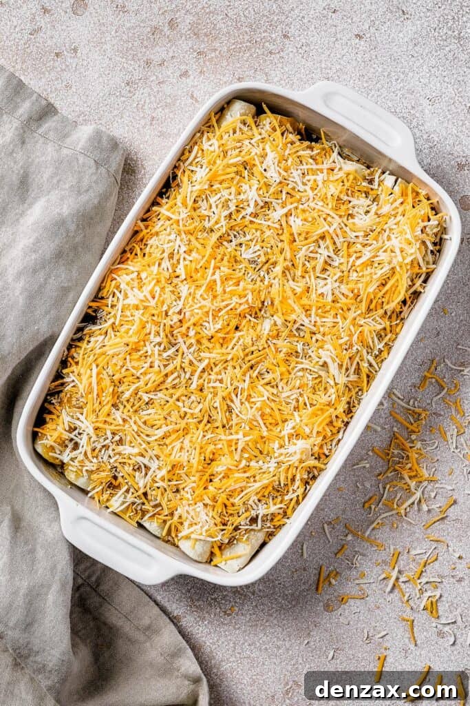 A white baking dish filled with neatly rolled chicken enchiladas, generously topped with a layer of shredded cheese and green salsa, ready for baking.