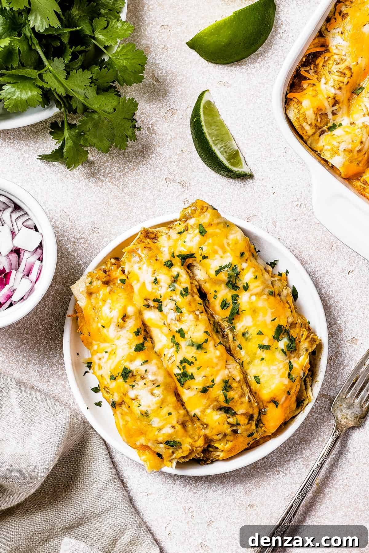 A single serving of chicken enchiladas on a small white plate, accompanied by small dishes of finely chopped red onion and fresh cilantro, ready for garnishing.