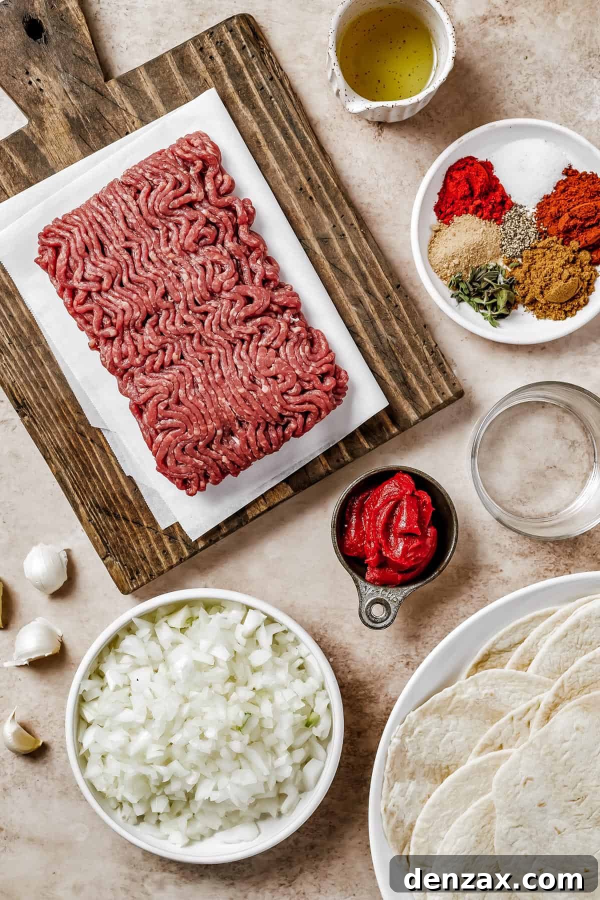 An overhead view of all the raw ingredients laid out: ground beef, cooking oil, various spices, water, tomato paste, chopped onions, and a stack of tortillas. This image clearly shows everything needed to make the tacos.