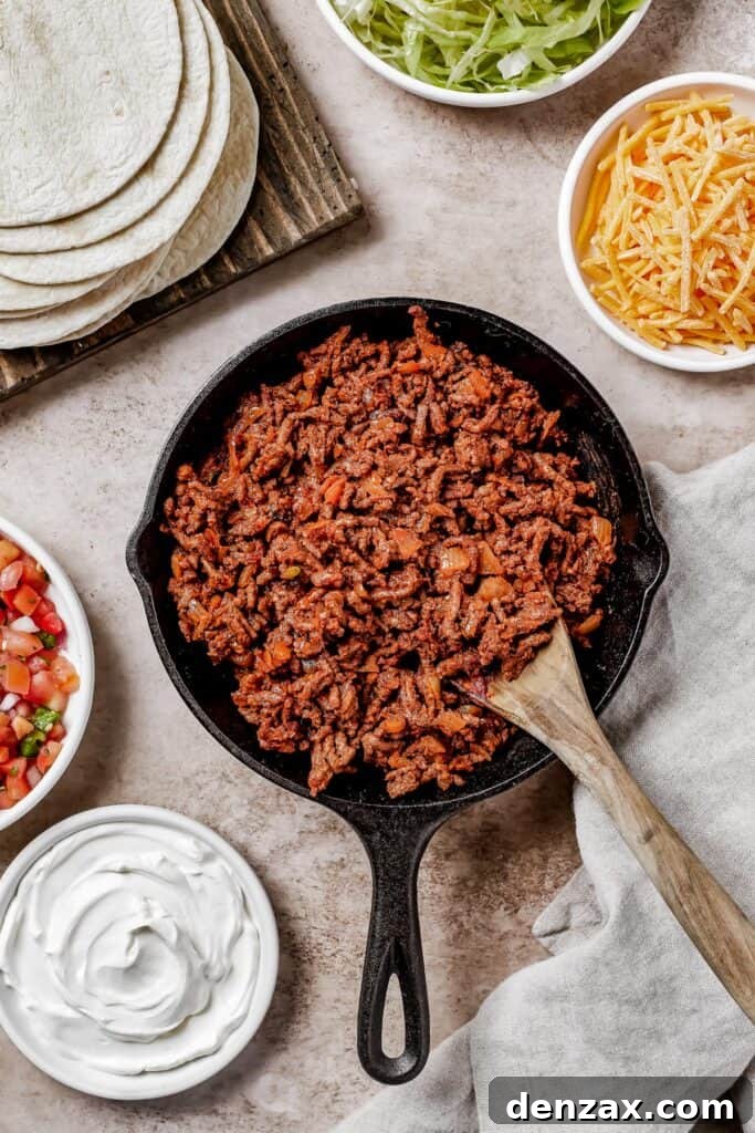 A wooden spoon stirring through a skillet full of richly seasoned cooked ground beef, ready to be served in tacos.