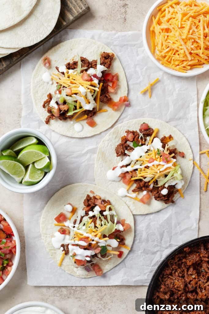 A vibrant spread of tortillas on a work surface, each generously topped with flavorful taco meat and a variety of colorful toppings, alongside bowls of lime wedges and shredded cheese.