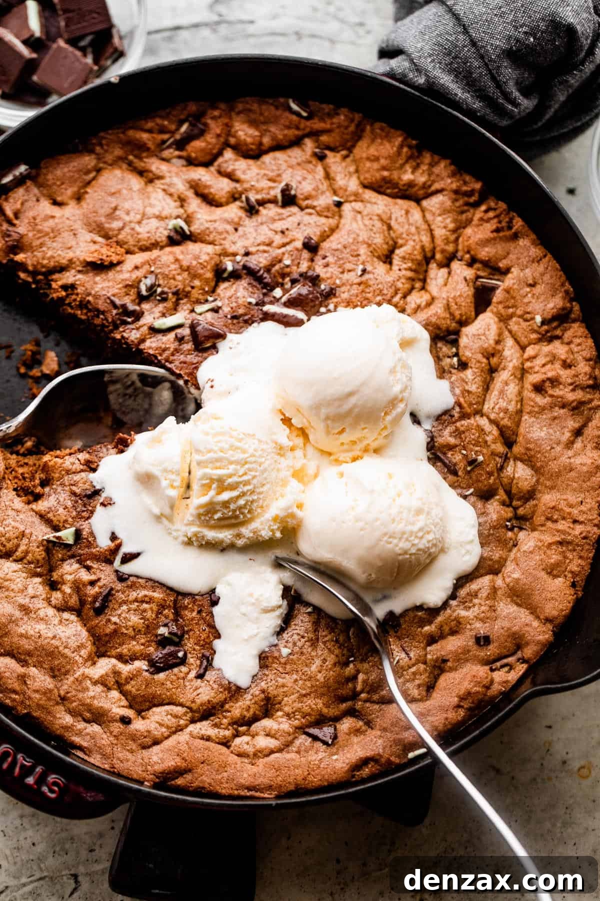 overhead shot of a skillet cookie inside the skillet, with three vanilla ice cream scoops on top and two large spoons digging into the cookie.