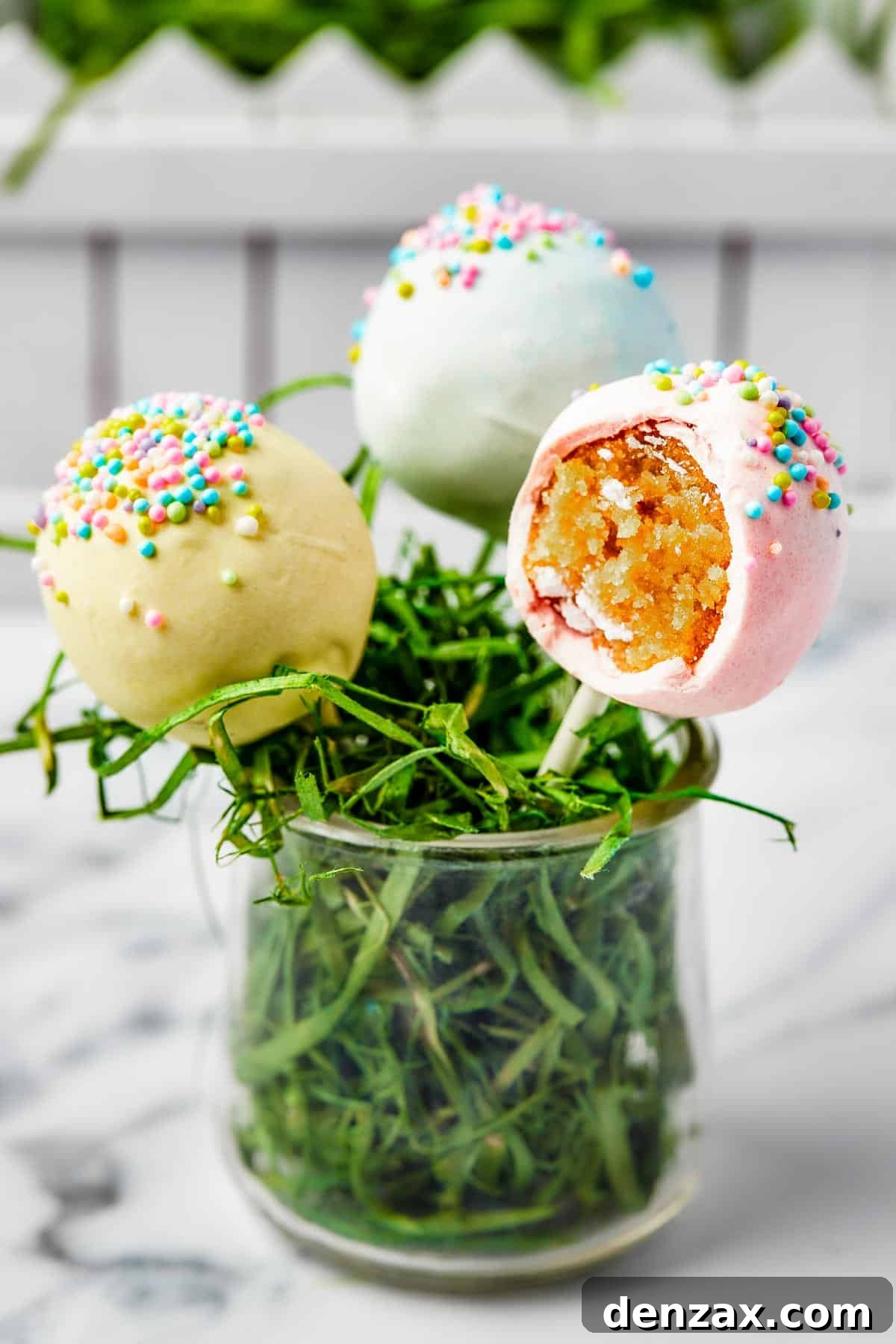 Three colorful cake lollipops nestled in a small glass filled with Easter grass, with one pop showing a delicious bite taken out.