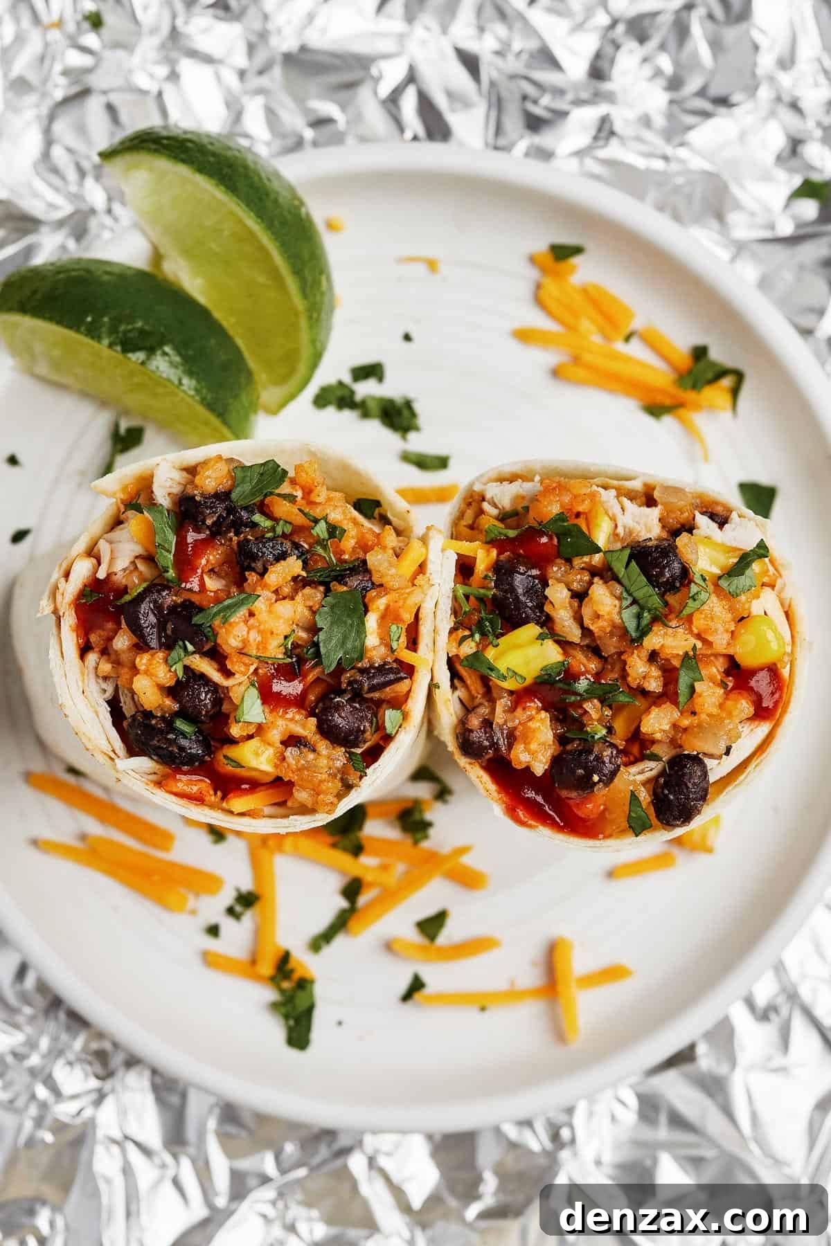 A beautifully presented plate featuring two halves of an Instant Pot Chicken Burrito, generously topped with shredded cheese and fresh lime wedges. In the background, aluminum foil-wrapped burritos and a bowl of extra cheese hint at a delicious spread.