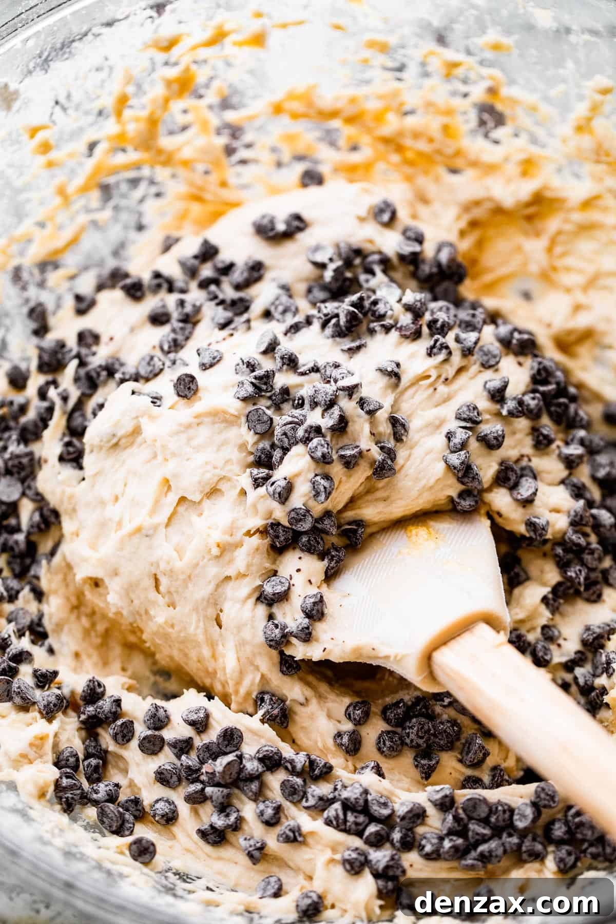 Close-up shot of mini chocolate chips being folded into a bowl of soft cookie batter with a rubber spatula.