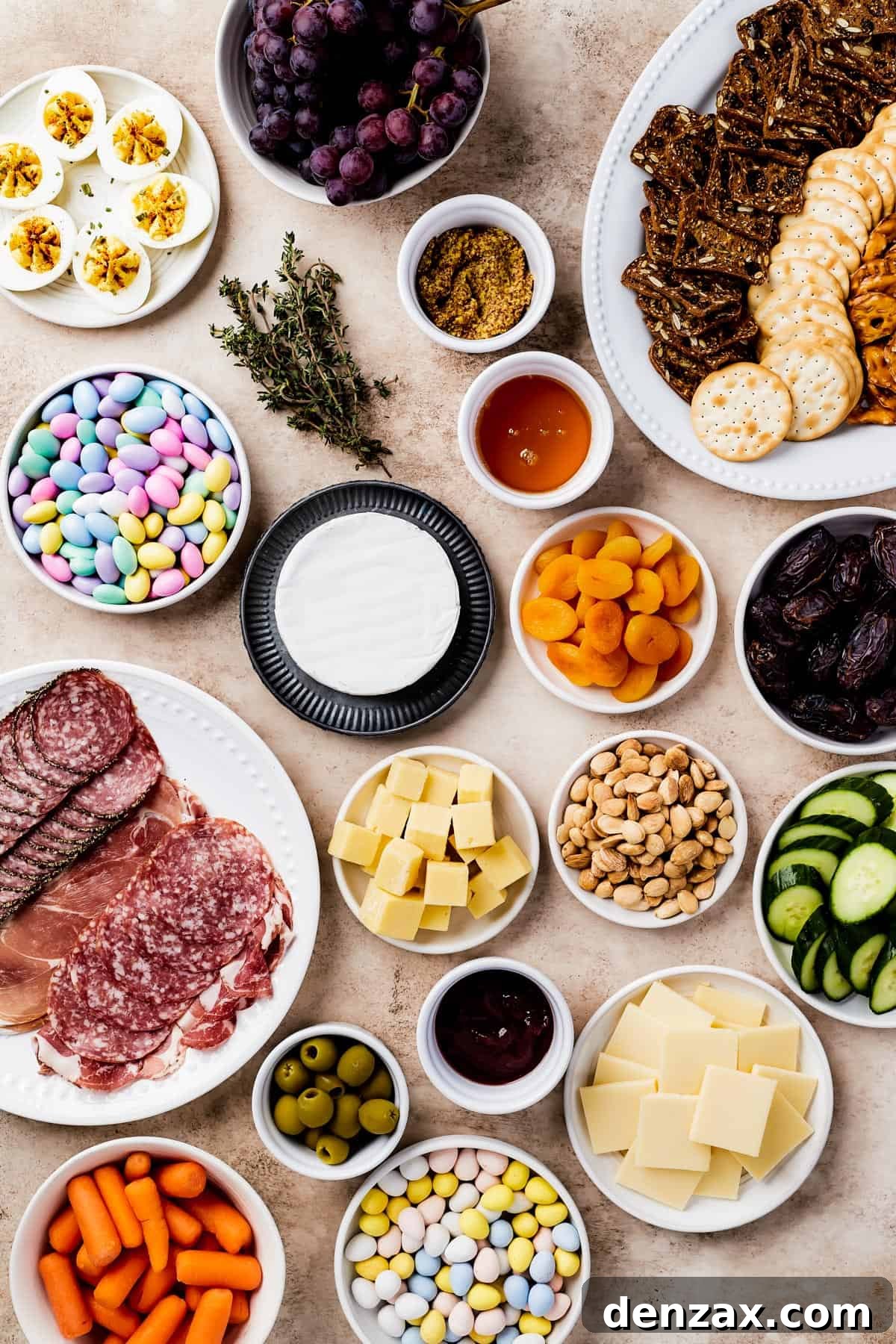 From top left: Deviled eggs, red grapes, a platter of assorted crackers, jordan almonds, fresh thyme, grainy mustard, strawberry preserves, a wheel of brie, dried apricots, medjool dates, a plate of sliced meats, cubed white cheddar, marcona almonds, sliced cucumber, castelvetrano olives, honey, gouda, all beautifully arranged for an Easter Charcuterie Board.