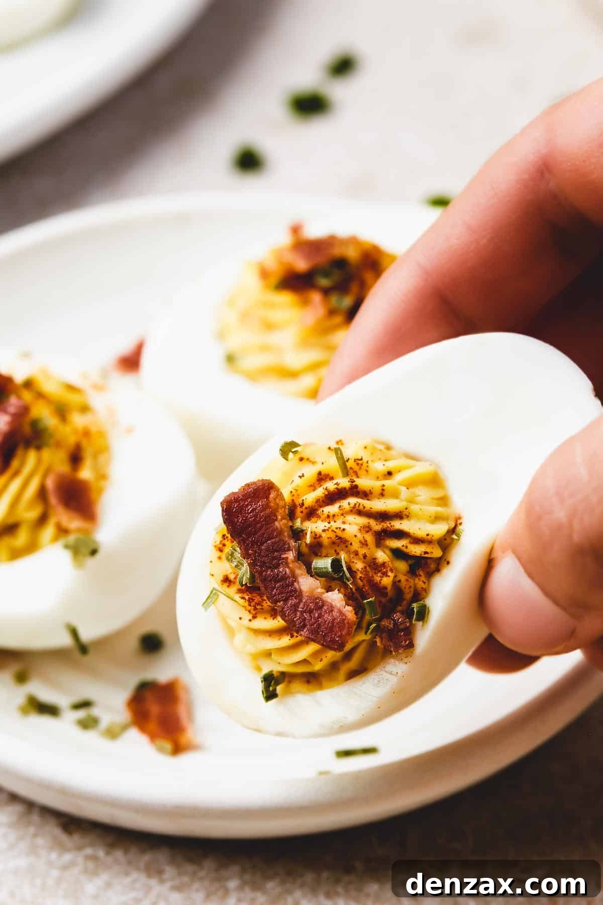 A woman's hand is gently lifting a beautifully piped deviled egg from a small serving plate, showcasing its fluffy filling and delicate garnish. The image captures the inviting and ready-to-eat nature of the appetizer.