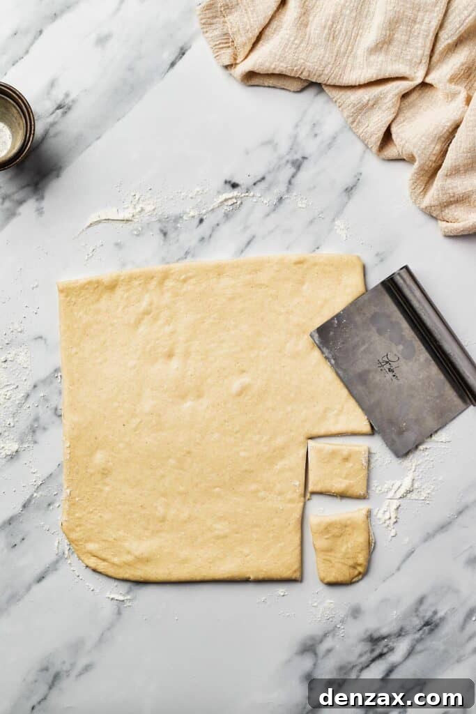 A section of rolled-out yeast dough on a marble surface, neatly pre-cut into small squares. A bench scraper rests nearby, ready for the next step.