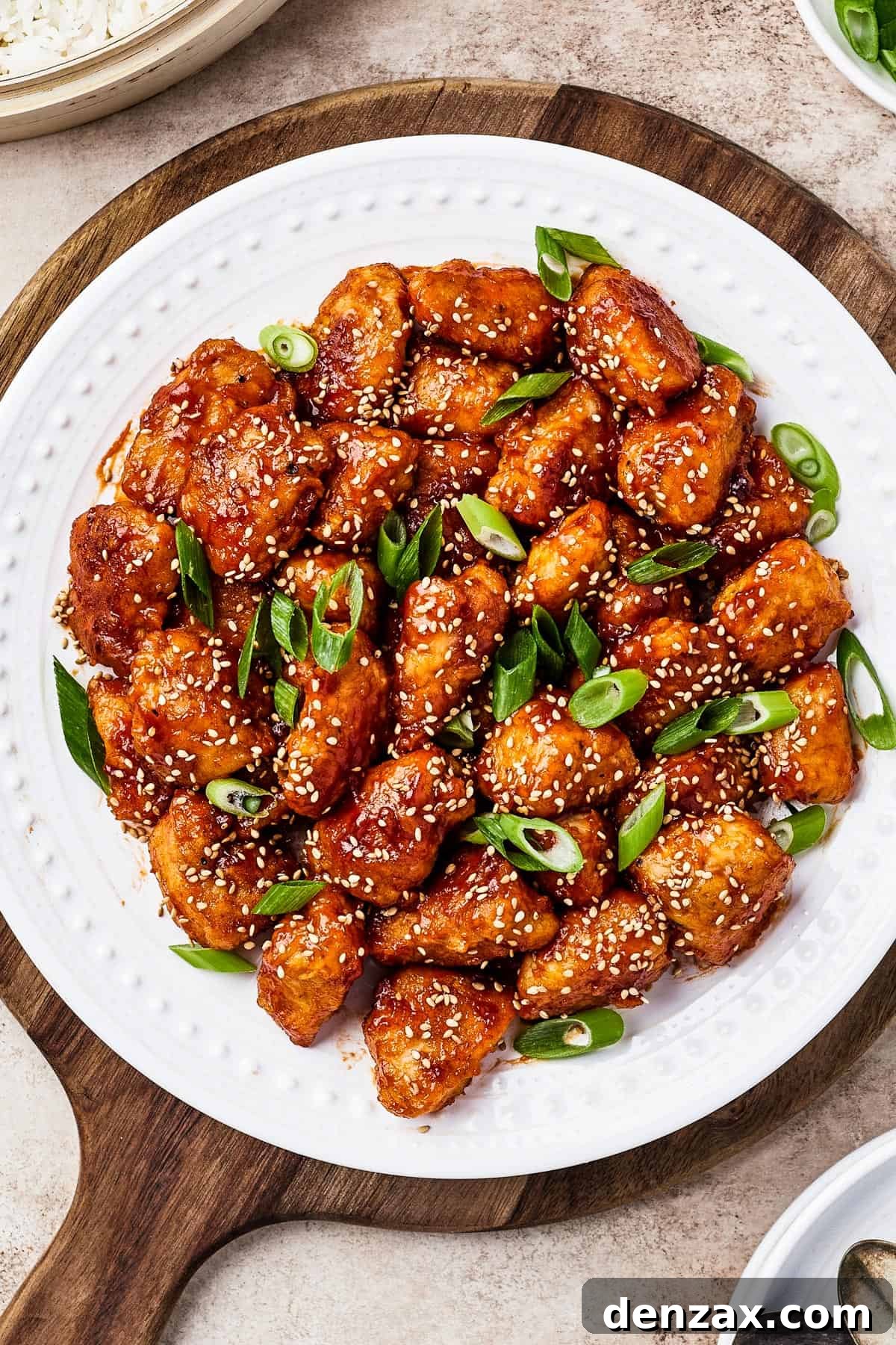 Overhead shot of homemade sweet and sour chicken served on a white plate with rice, garnished with fresh green onions and toasted sesame seeds.
