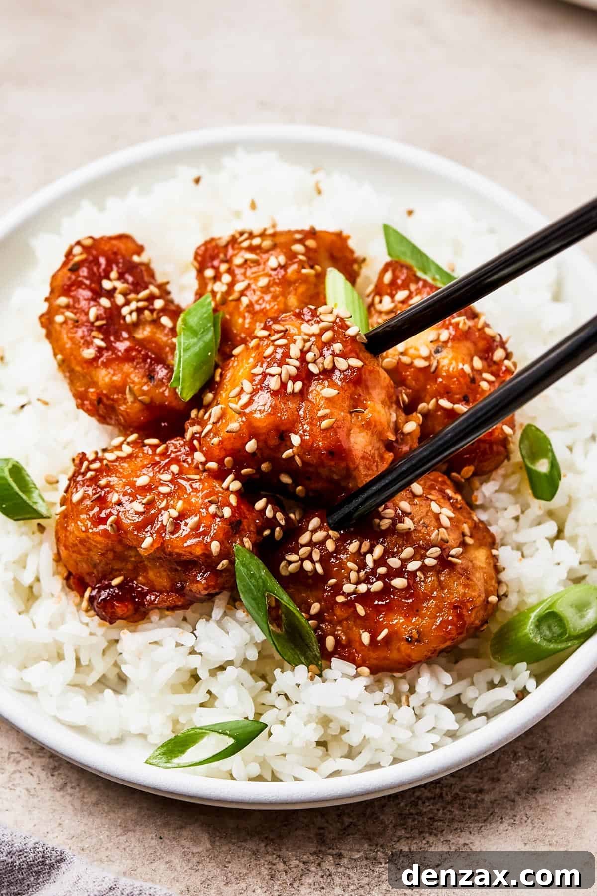 A small plate of perfectly cooked homemade sweet and sour chicken served over a bed of white rice, with chopsticks holding one piece of saucy chicken.