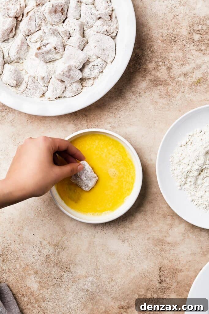 A chicken breading station set up with breaded chicken pieces, an egg wash bowl, a bowl of flour, and more floured chicken on a plate.