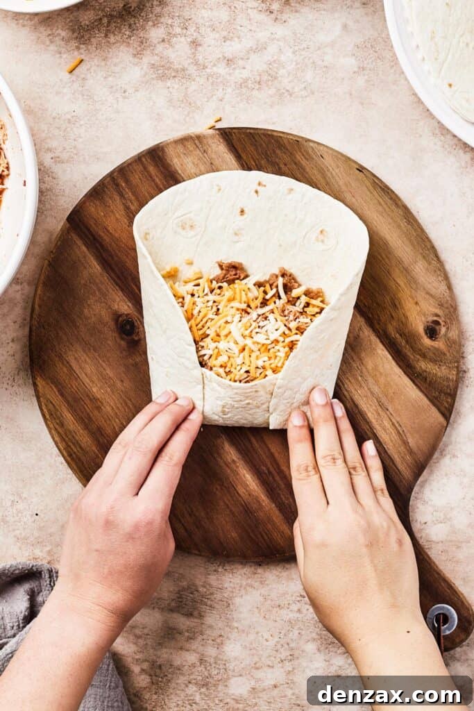 A person's hands skillfully folding the bottom and sides of a flour tortilla over the filling, demonstrating the rolling technique for a chimichanga.