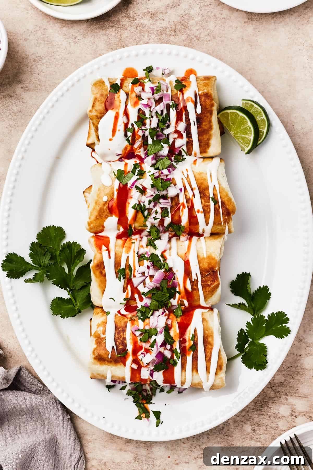A vibrant plate of fried chicken chimichangas beautifully garnished with swirls of sour cream, splashes of hot sauce, fresh cilantro, lime wedges, and finely chopped red onion.