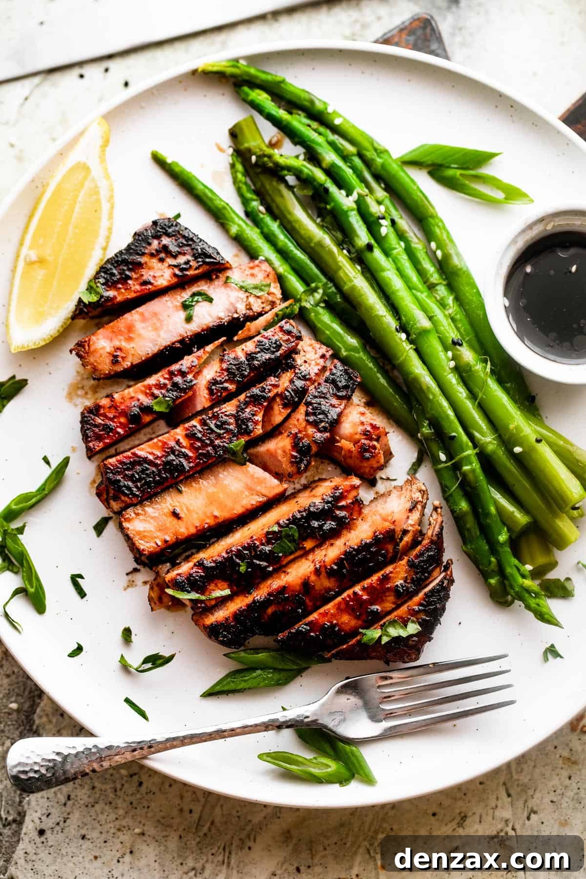 Enjoy a flavorful meal with this easy recipe for Blackened Balsamic Tuna Steaks, featuring a perfect blend of spicy Cajun spices and sweet balsamic vinaigrette. Wide overhead shot of a white dinner plate with a sliced blackened balsamic tuna steak, fresh asparagus stalks, a side of balsamic dipping sauce, and a bright lemon wedge.