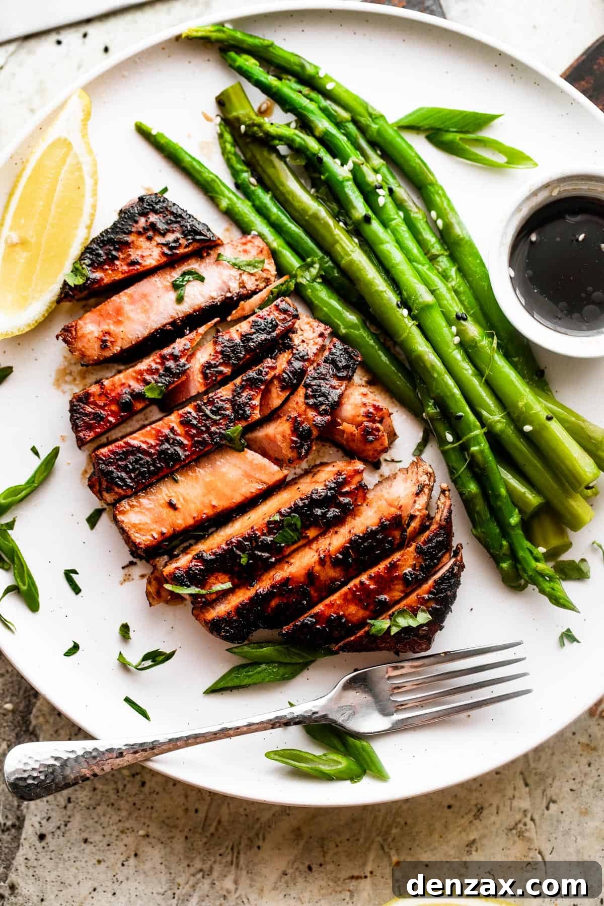 Enjoy a flavorful meal with this easy recipe for Blackened Balsamic Tuna Steaks, featuring a perfect blend of spicy Cajun spices and sweet balsamic vinaigrette. Overhead shot of a sliced blackened balsamic tuna steak served on a white dinner plate with asparagus stalks, a small bowl of dipping sauce, and lemon slices.
