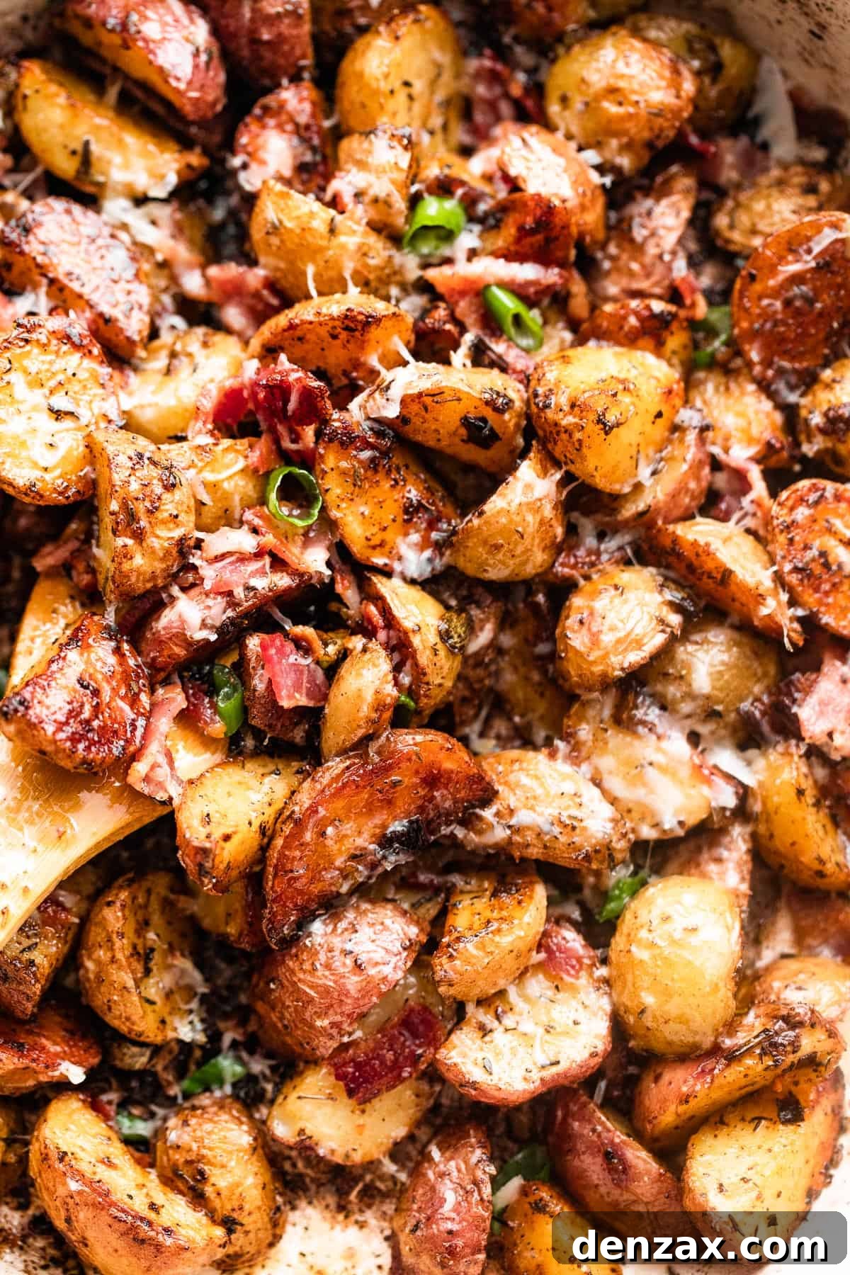 close up shot of a wooden spoon stirring through roasted potatoes with bacon and cheese, showing golden potatoes and melted cheese.