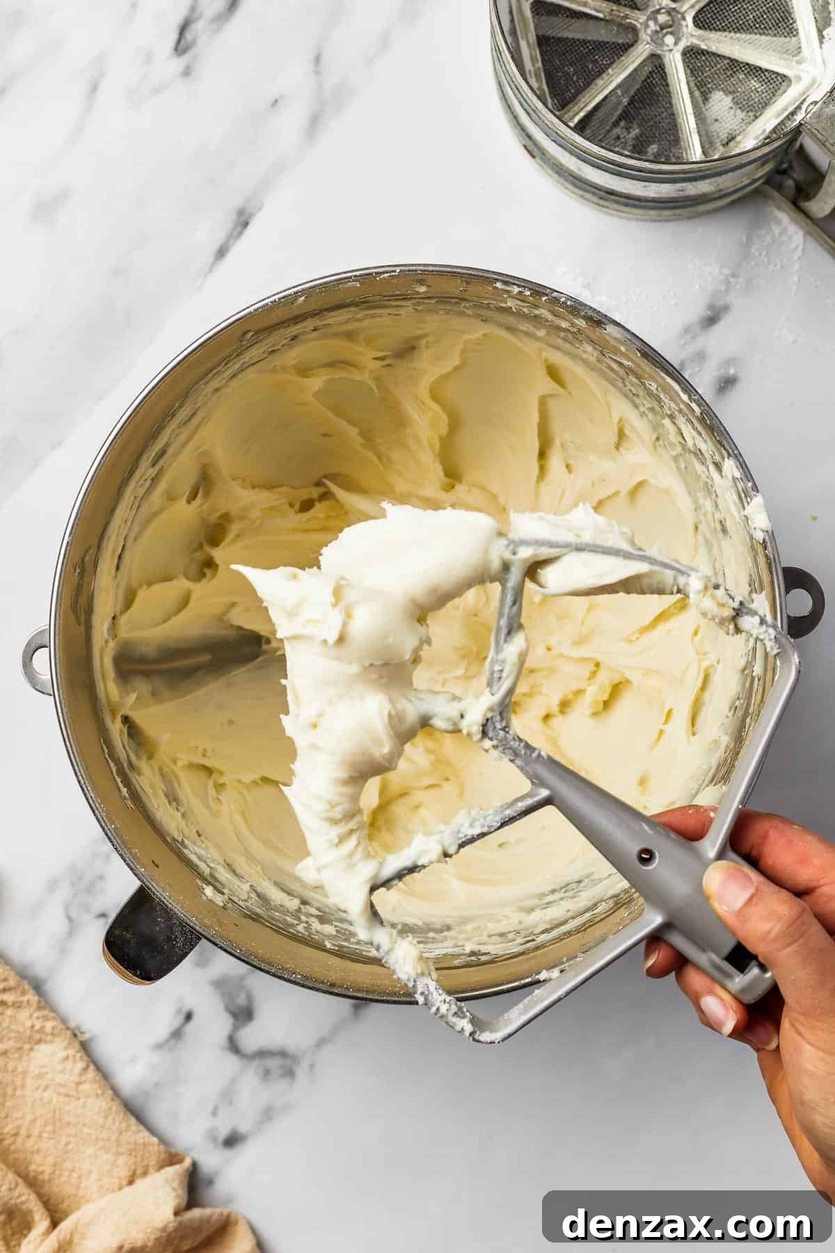 A woman's hand gently lifts a paddle beater from a metal mixing bowl, revealing perfectly whipped, creamy white cream cheese frosting, ready for baking.