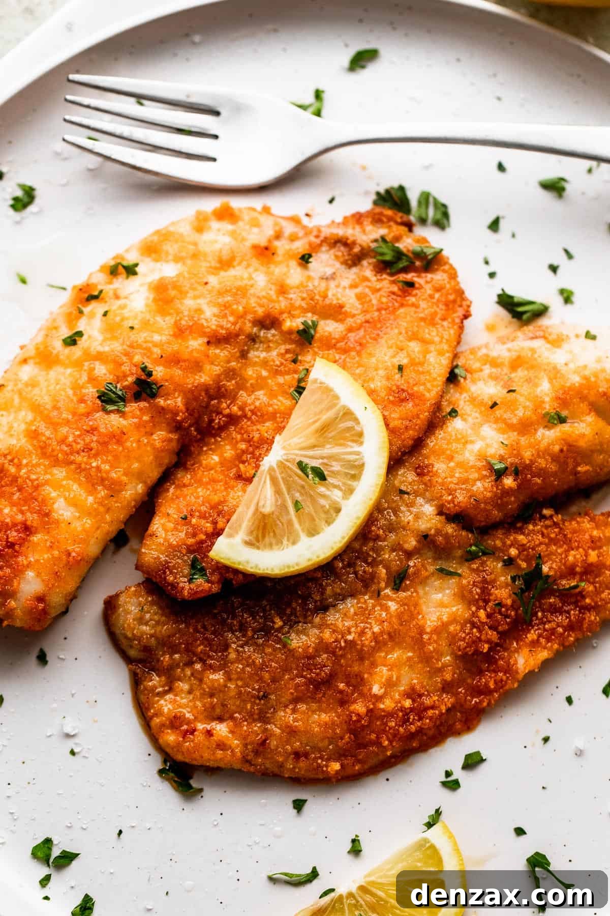 two cooked tilapia fillets placed on a white plate and topped with a slice of lemon.