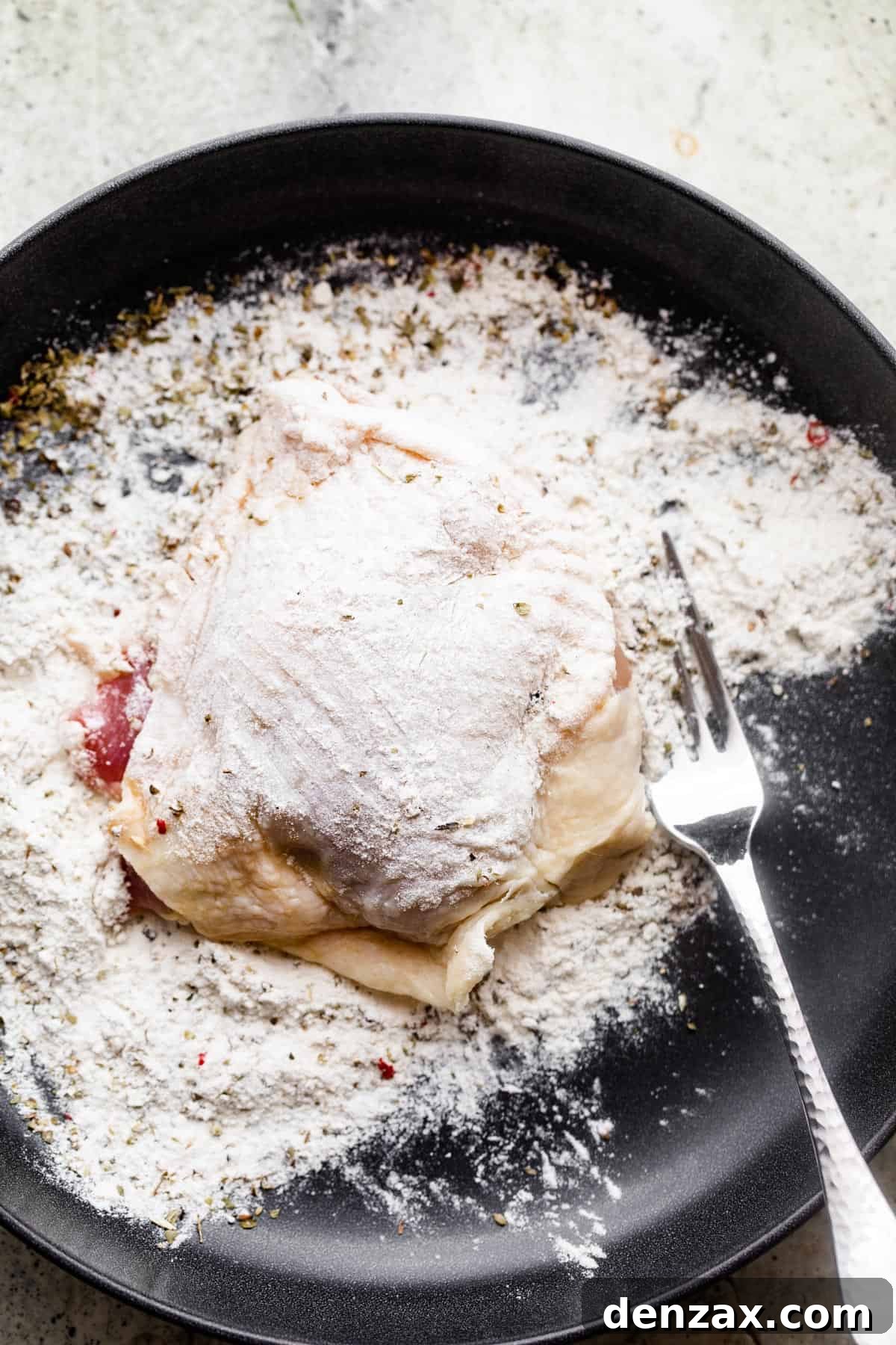 Raw chicken thigh set on a black plate filled with flour, and a fork set right next to the chicken thigh.