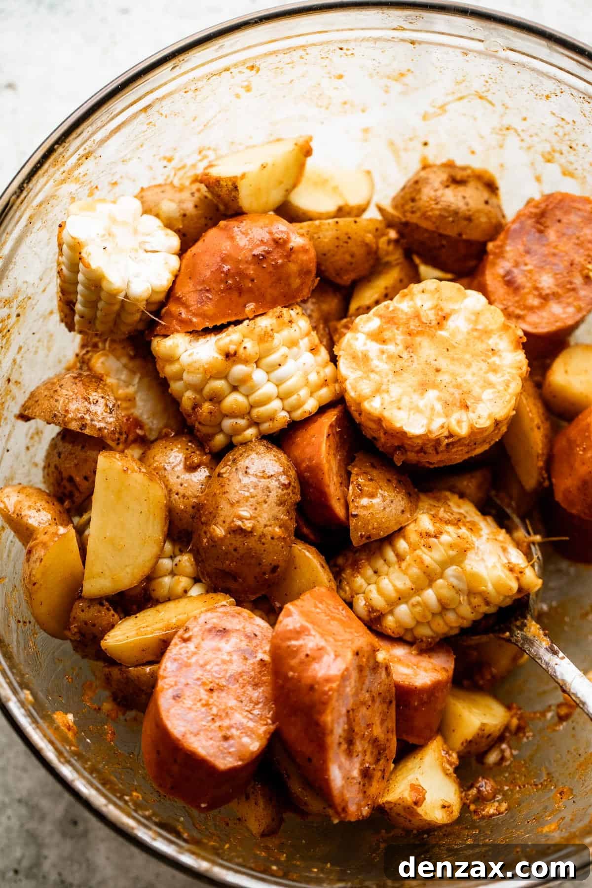 Overhead shot of perfectly prepped ingredients: sliced new potatoes, corn on the cob rounds, andouille sausage slices, and raw jumbo shrimp, all ready in a glass mixing bowl.
