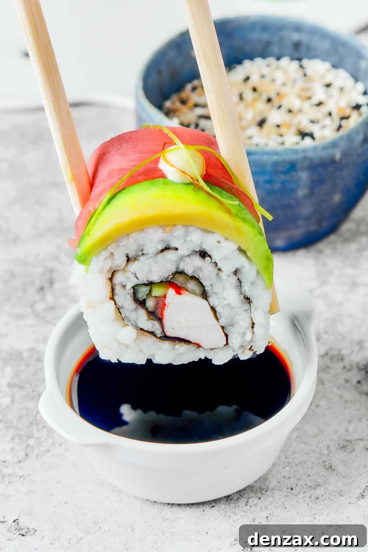 A bite of sushi held in a pair of chopsticks. A small plate of sushi and a container of soy sauce is in the background.