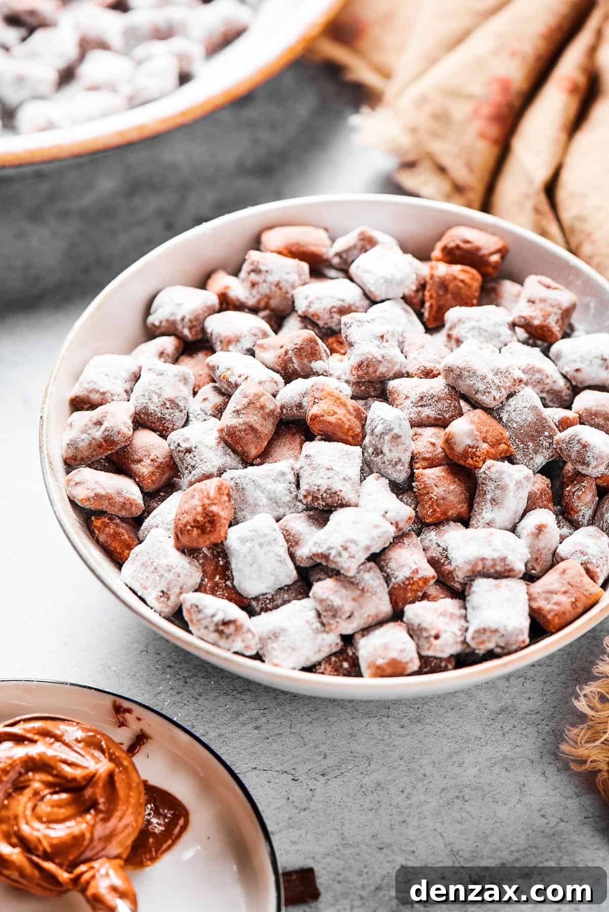 Puppy Chow 12 A shallow bowl filled with candy-coated cereal, near a beige napkin. Peanut butter and chocolate chunks are nearby on the table.