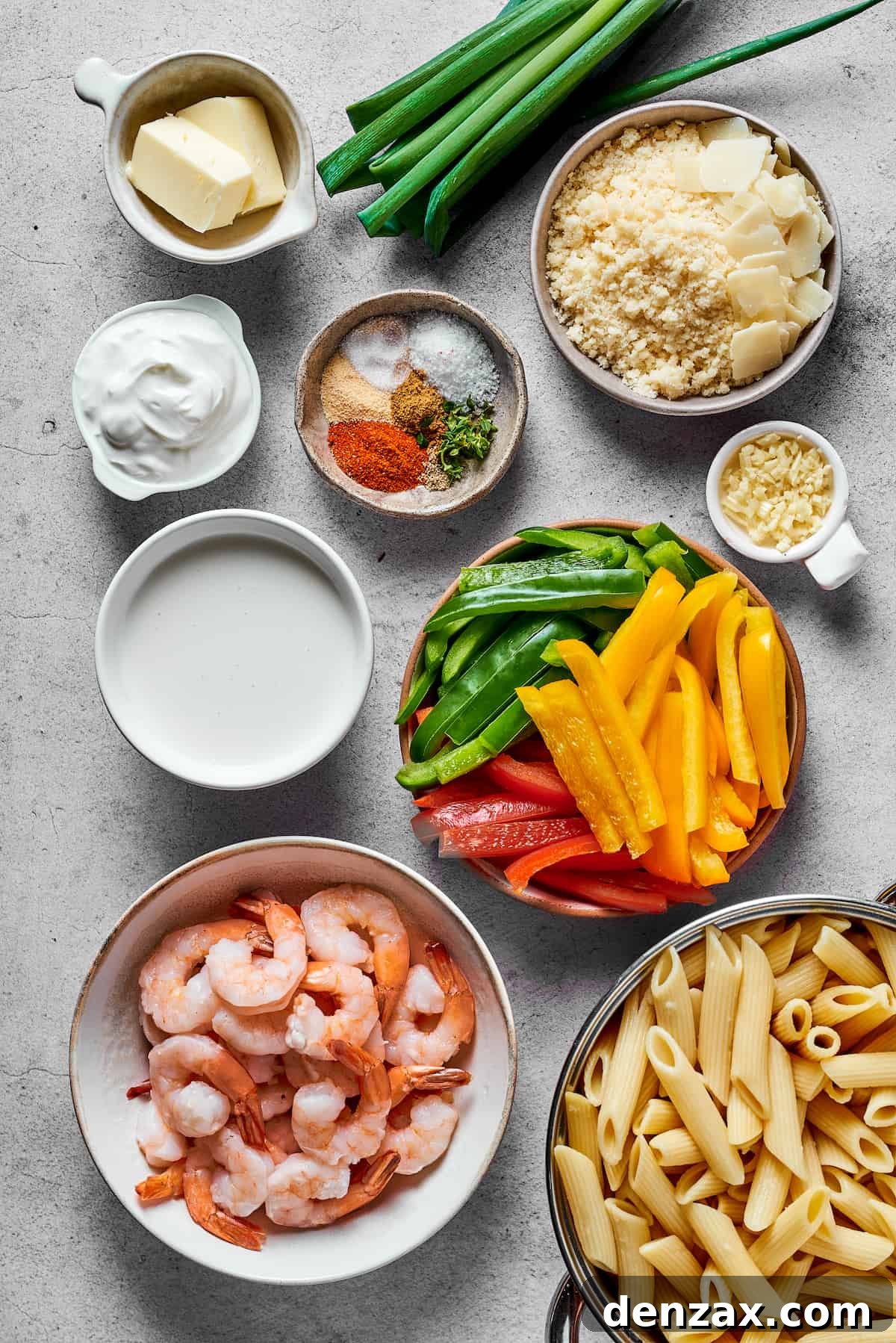An array of Rasta Pasta ingredients including butter, green onions, parmesan, cream cheese, spices, garlic, heavy cream, sliced bell peppers, shrimp, and penne pasta.