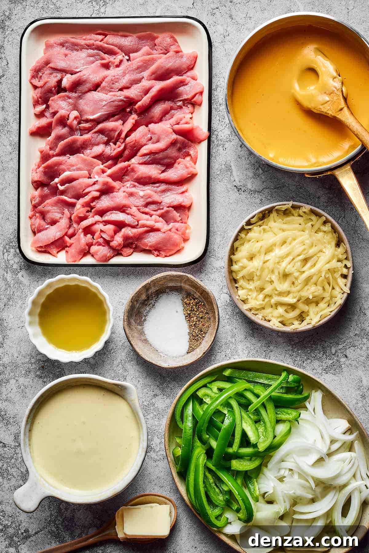 All the ingredients for Philly Cheesesteak Burgers laid out: thinly sliced raw steak, a bowl of Cheez Whiz, olive oil, salt and pepper shakers, grated Provolone cheese, a jar of aioli, sliced onions, sliced green bell peppers, and a stick of butter.