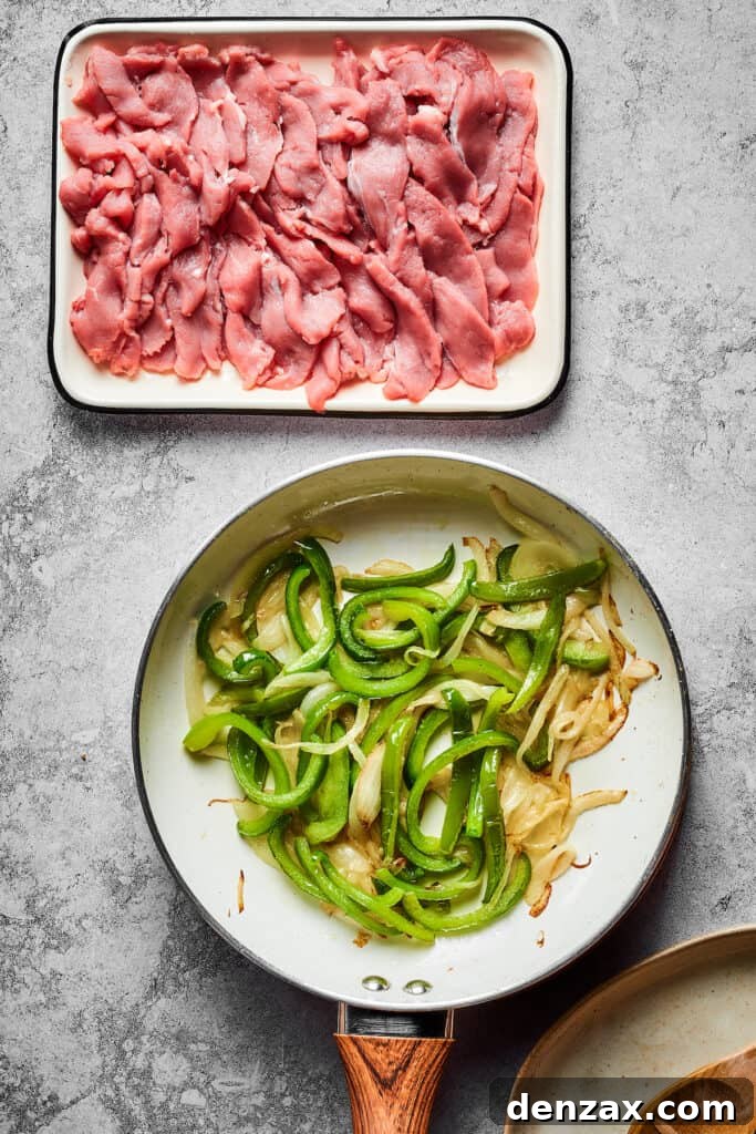 A non-stick skillet contains a colorful mix of sautéing onions and green bell peppers, while a white enamel dish nearby holds raw, thinly sliced steak, ready for cooking.