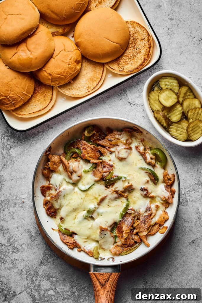 A culinary scene: toasted sandwich buns and a small dish of dill pickle slices sit beside a skillet overflowing with cooked steak, onions, and bell peppers, all topped with perfectly melted Provolone cheese.