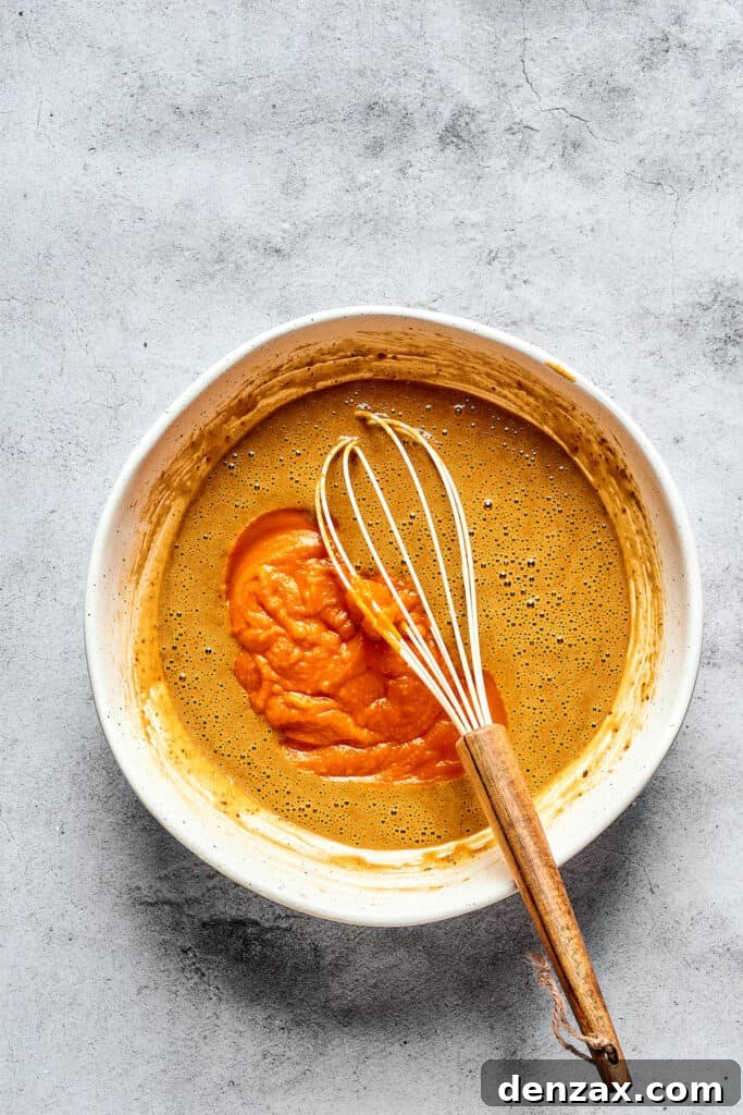 Wet muffin batter ingredients in a mixing bowl with a whisk, including pumpkin puree.