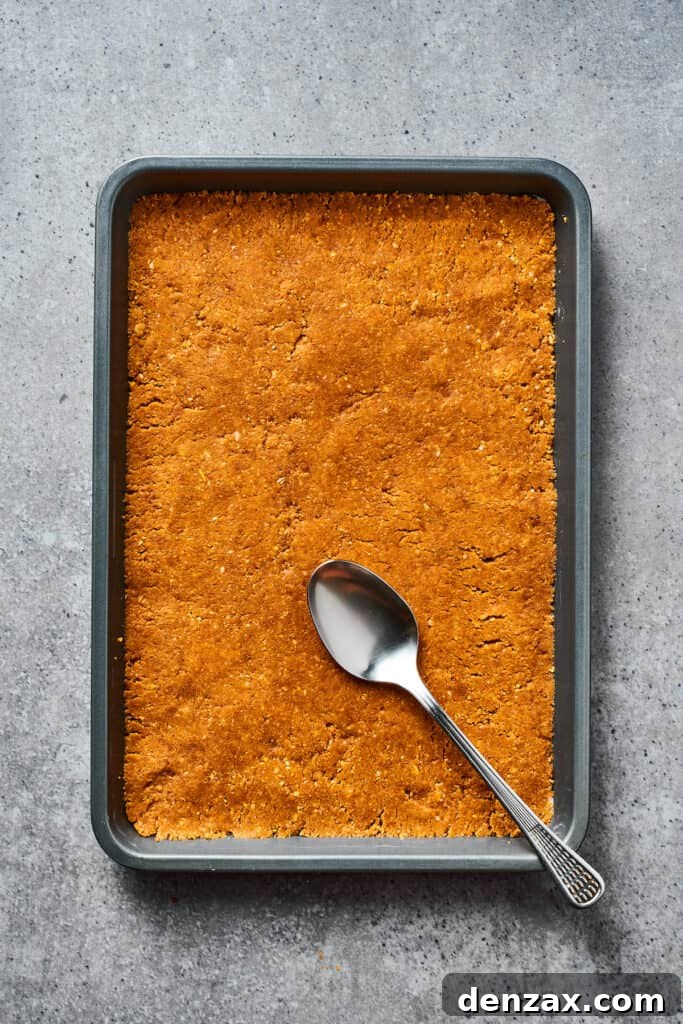 A cookie crust pressed into a parchment-lined baking dish with a spoon, ready for the filling.