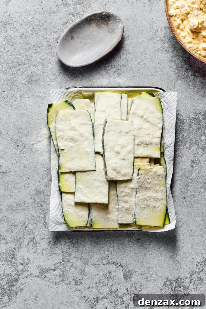 Thin strips of raw zucchini, delicately coated in flour, resting on a paper-towel-lined tray, ready for layering.