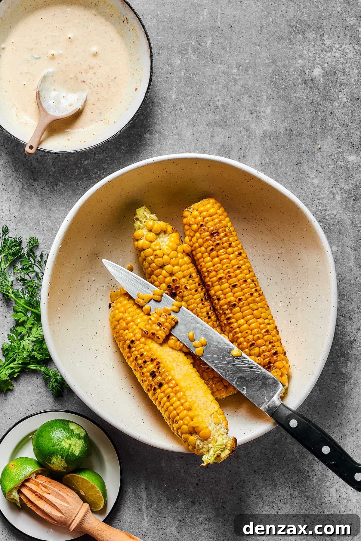 Grilled corn on the cob with some kernels already cut into a bowl, and a knife resting nearby, indicating the next step.